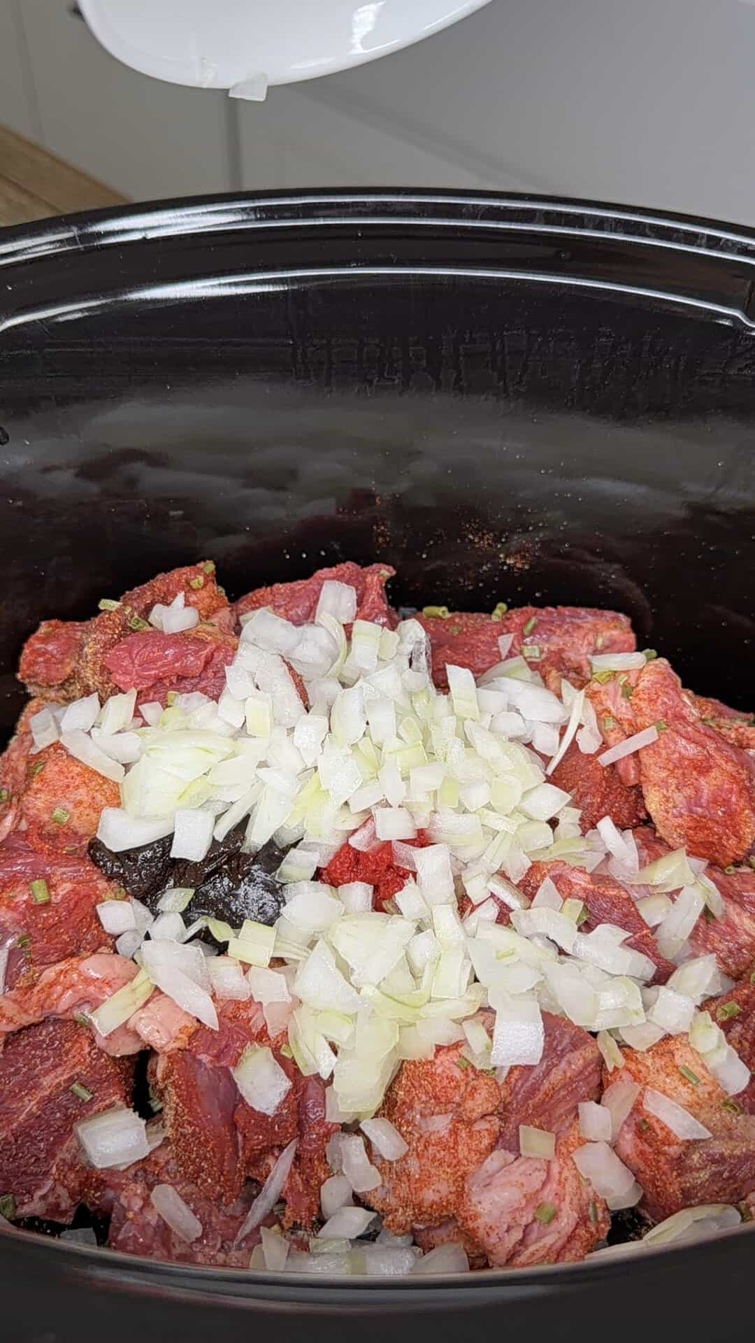 Diced onions and seasoned raw beef chunks in a black slow cooker, ready to be cooked. A white bowl is partially visible above, as if just used to add the onions.