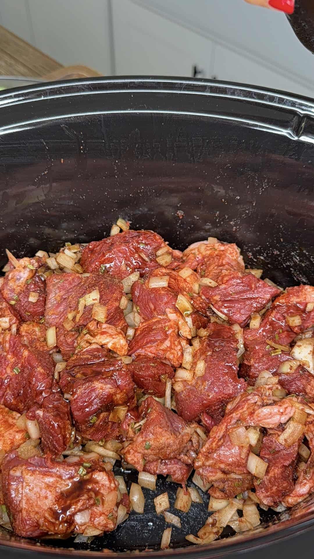 Chunks of raw beef mixed with chopped onions and seasonings inside a black slow cooker, ready to be cooked.