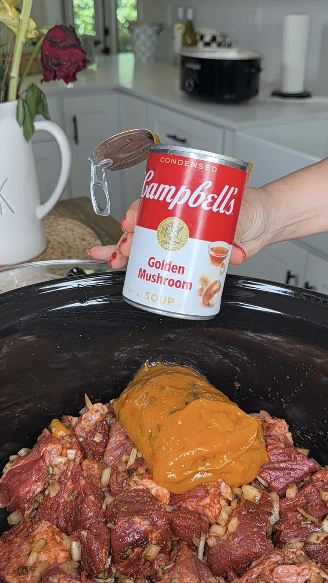 A hand pours Campbell’s Golden Mushroom soup from a can into a slow cooker filled with seasoned beef chunks and chopped onions in a kitchen setting.