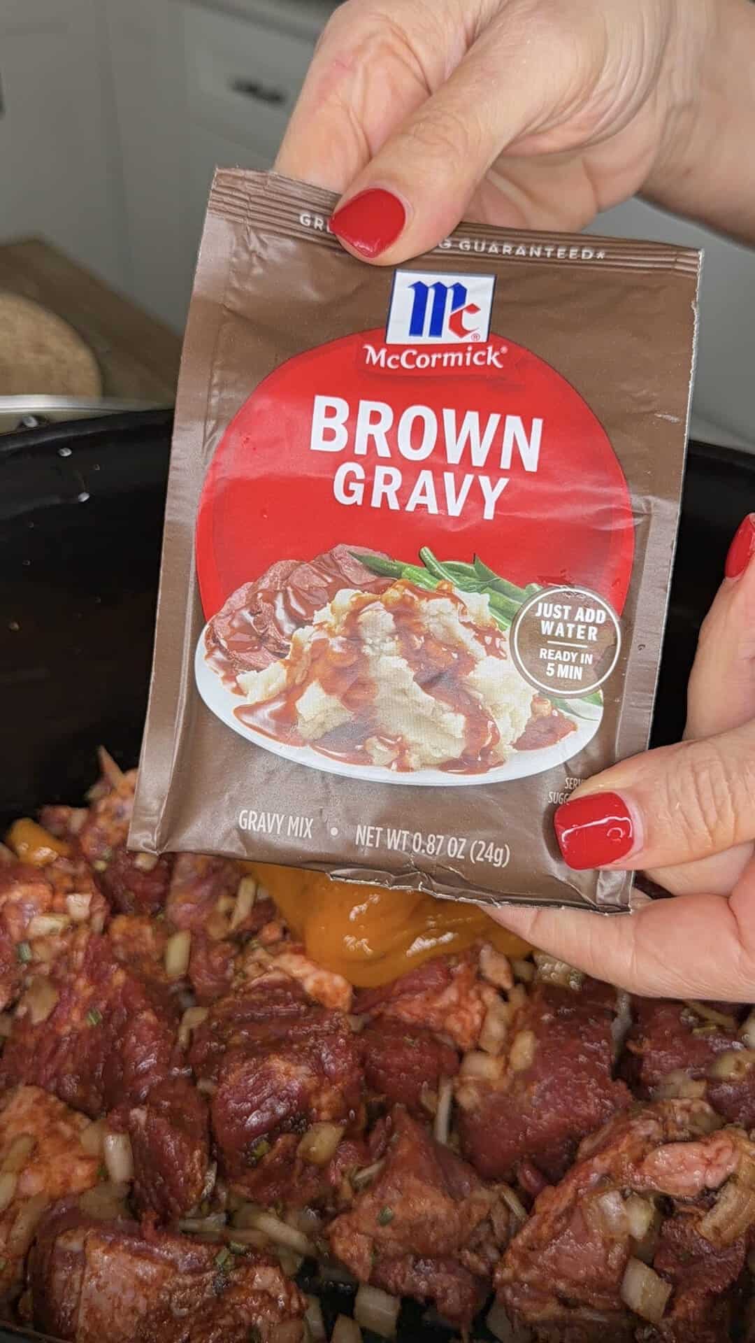 A hand with red nails holds a McCormick Brown Gravy packet over a slow cooker filled with seasoned beef and chopped onions.