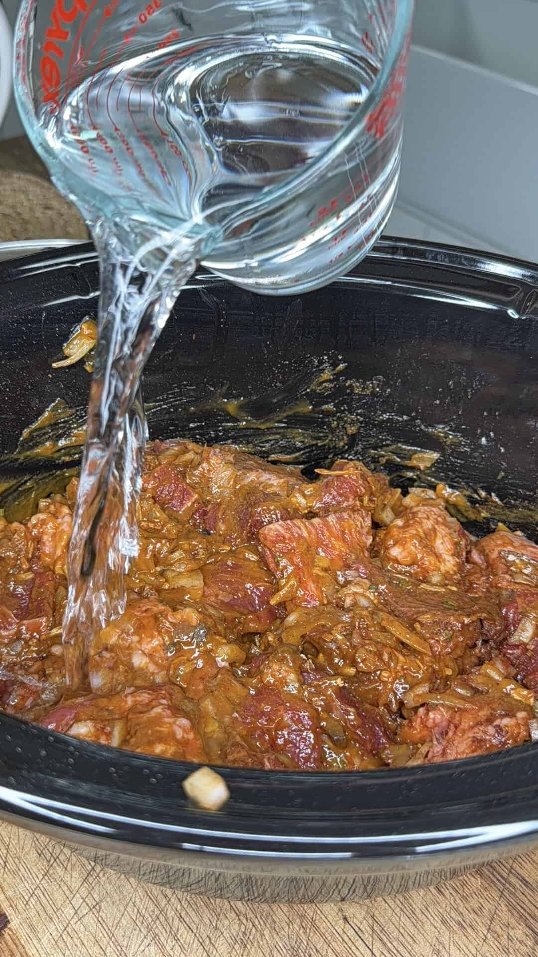 Water is being poured from a glass measuring cup into a slow cooker filled with seasoned meat and chopped onions.