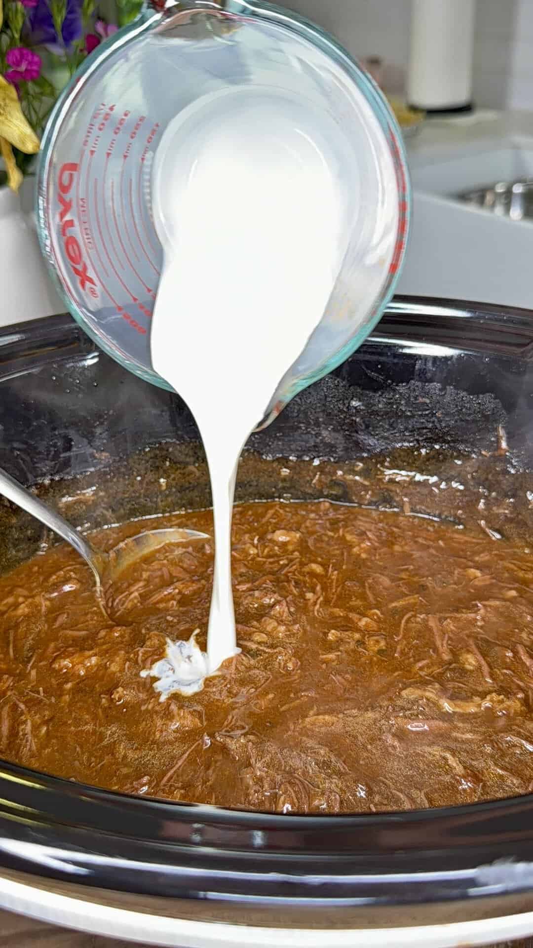 A glass measuring cup pours a creamy white liquid into a slow cooker filled with shredded meat in a brown sauce. A metal spoon rests in the cooker.