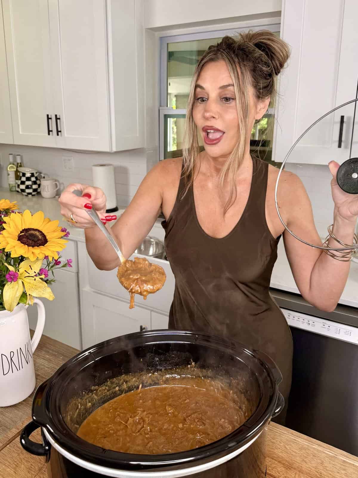 A woman in a brown tank top holds a slow cooker lid and ladles stew from a slow cooker in a bright kitchen. Sunflowers in a vase and a “DRINK” jug sit on the wooden counter nearby.