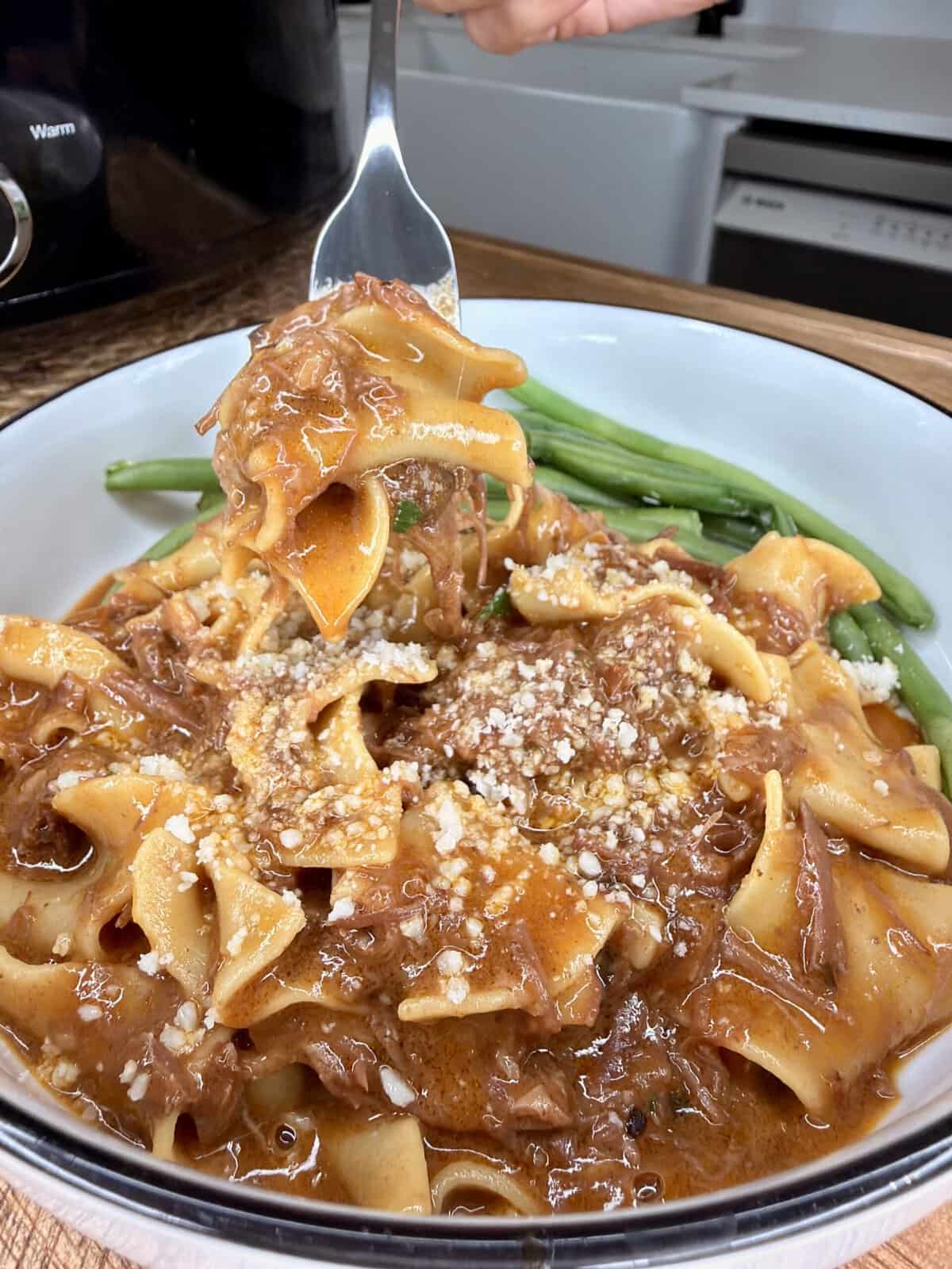 Wide egg noodles in a rich brown beef sauce, topped with grated cheese, served with green beans on the side. A fork lifts a portion of noodles from the bowl.