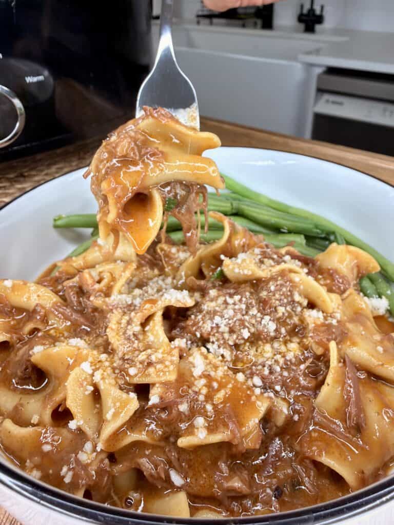 A bowl of pasta with a rich, reddish meat sauce, topped with grated cheese, with green beans on the side. A fork lifts a bite of pasta above the bowl. A kitchen counter and appliances are in the background.