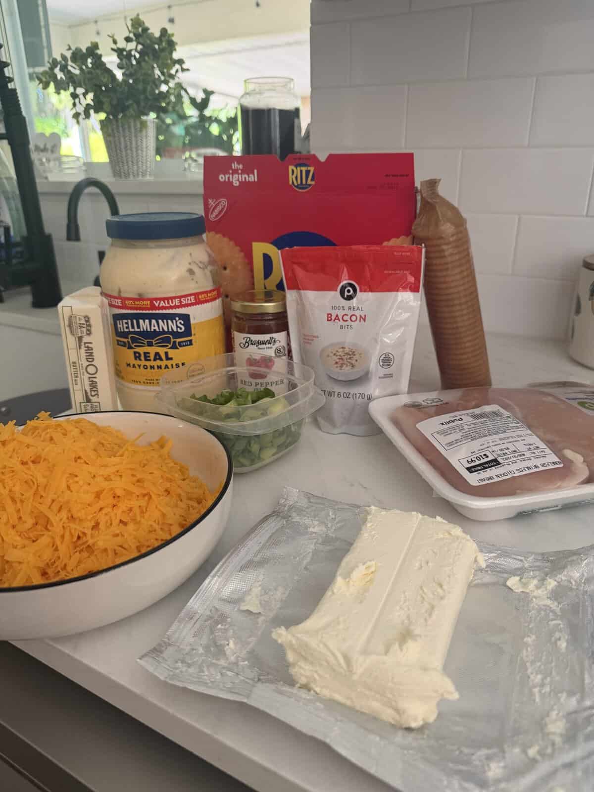 Ingredients on a kitchen counter: shredded cheddar, cream cheese, green onions, mayo, Ritz crackers, cooked bacon, ham, and a spice container, with plants and a window in the background.