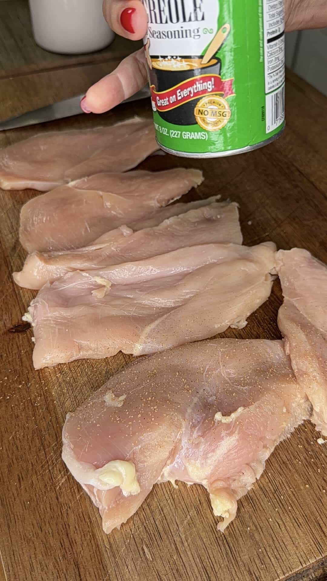 Raw chicken breasts on a wooden cutting board being seasoned with a green can of Creole seasoning. A hand with red-painted nails holds the can above the chicken slices.