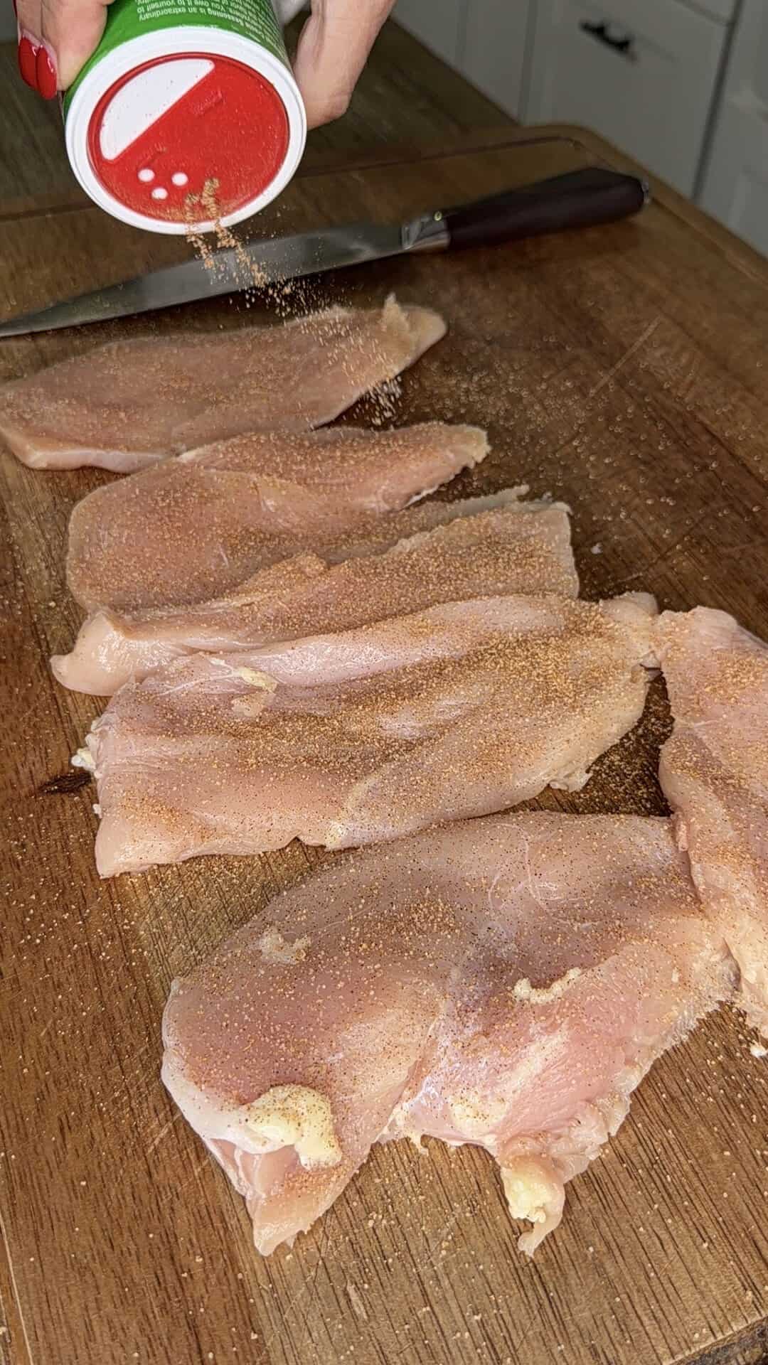 A hand sprinkles seasoning from a green container onto raw chicken breasts arranged on a wooden cutting board, with a knife in the background.