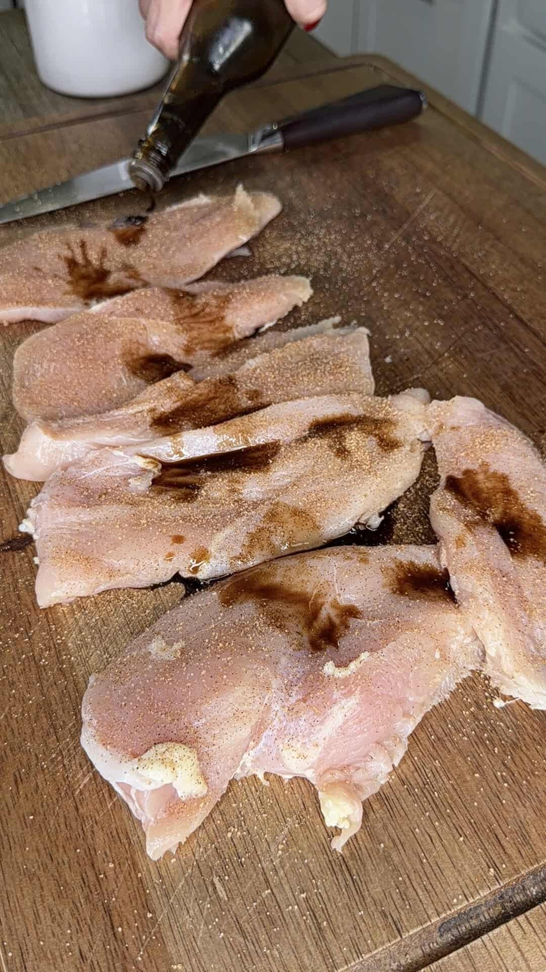 Raw chicken breasts on a wooden cutting board are being seasoned and drizzled with a dark liquid, possibly soy sauce. A knife is visible in the background.