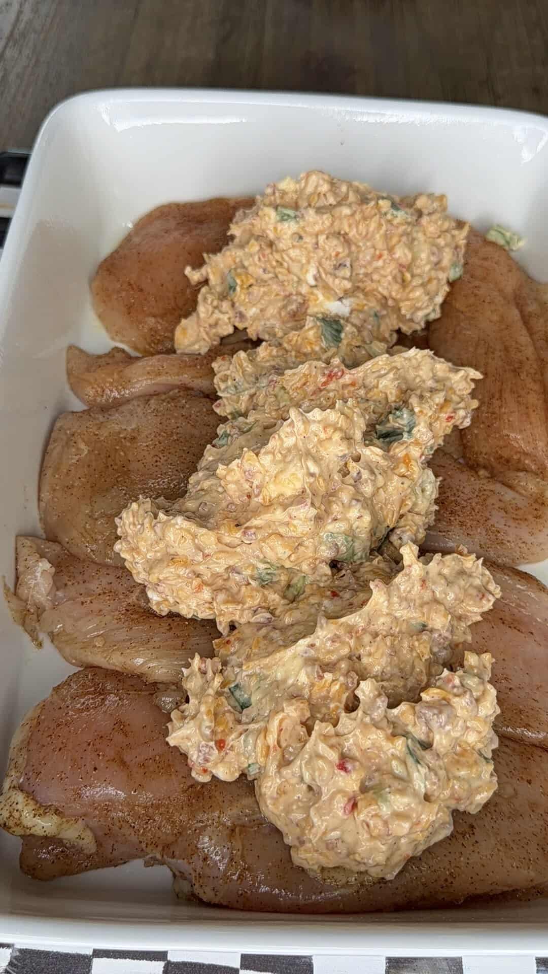 Raw chicken breasts in a baking dish, each topped with a mixture of shredded cheese, chopped vegetables, and a creamy, seasoned filling.