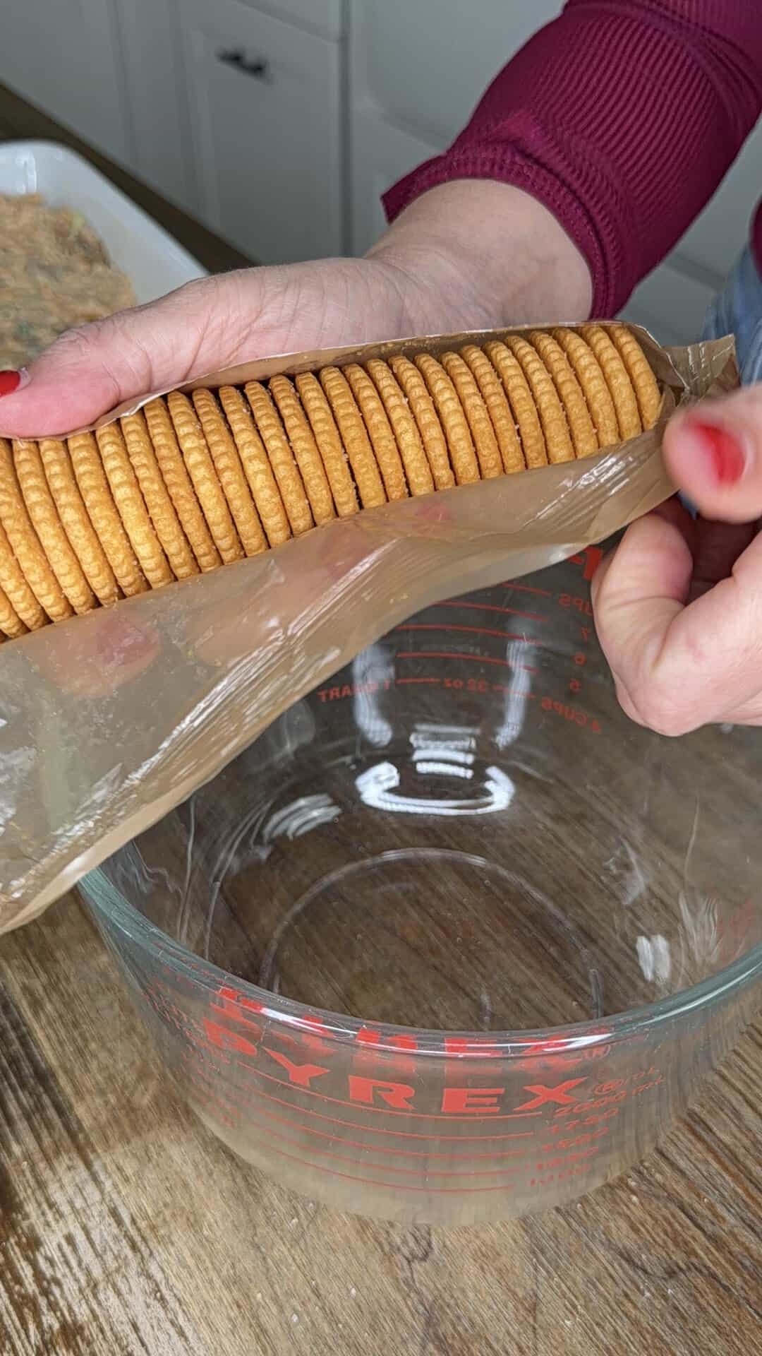 A person holding a package of round golden crackers over a clear glass Pyrex mixing bowl in a kitchen.