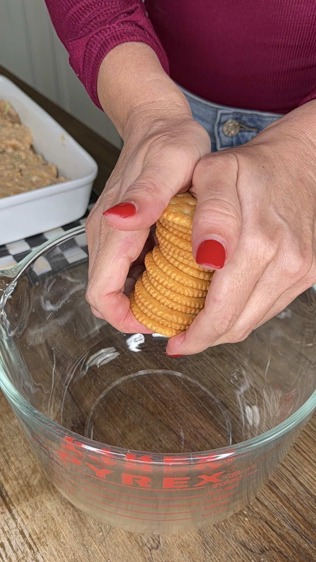A person with red nail polish crushes a stack of round crackers over a large glass Pyrex mixing bowl on a wooden surface.