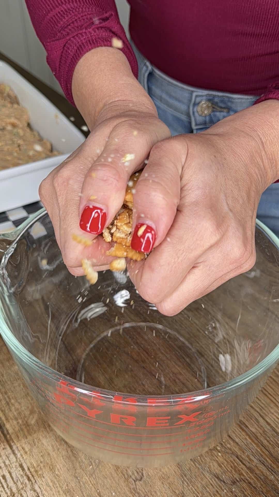 A person with red painted nails squeezes soaked bread over a glass measuring cup, with a baking dish and bread visible in the background.