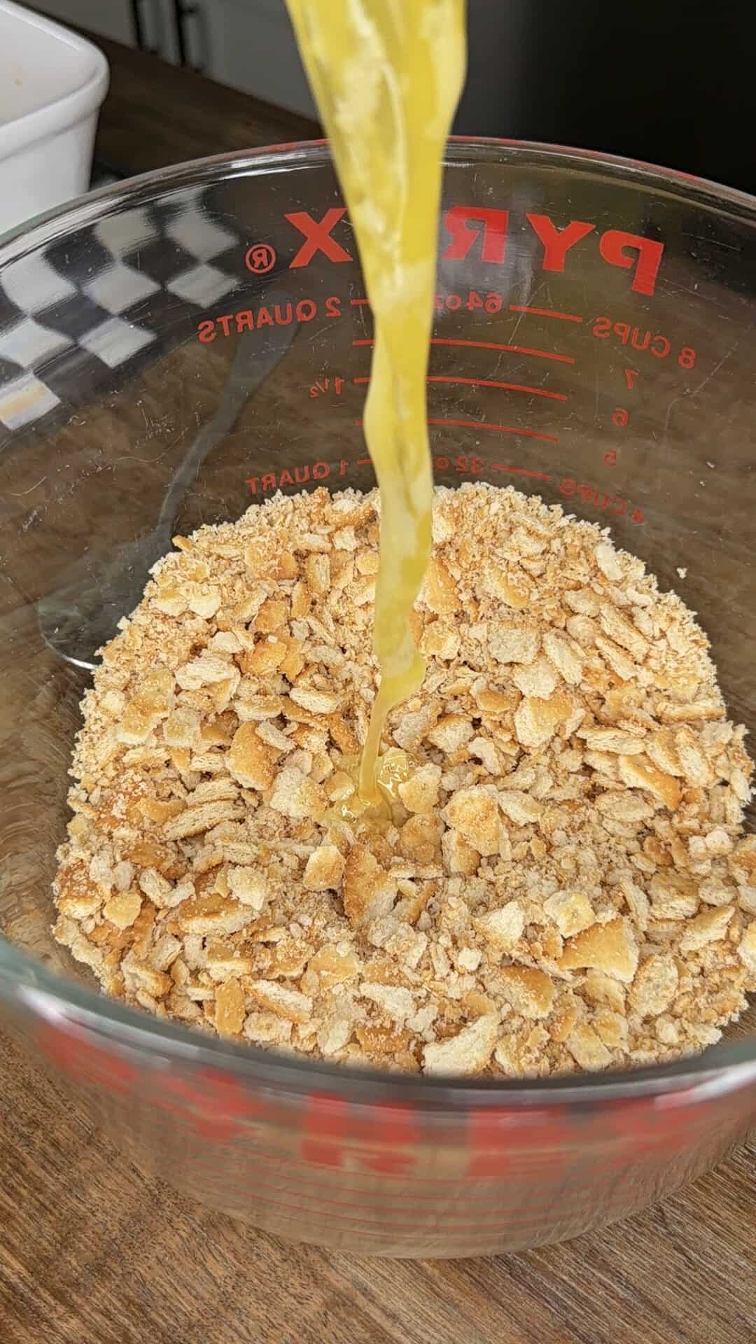 Melted butter being poured into a glass measuring cup filled with crushed crackers on a wooden surface.