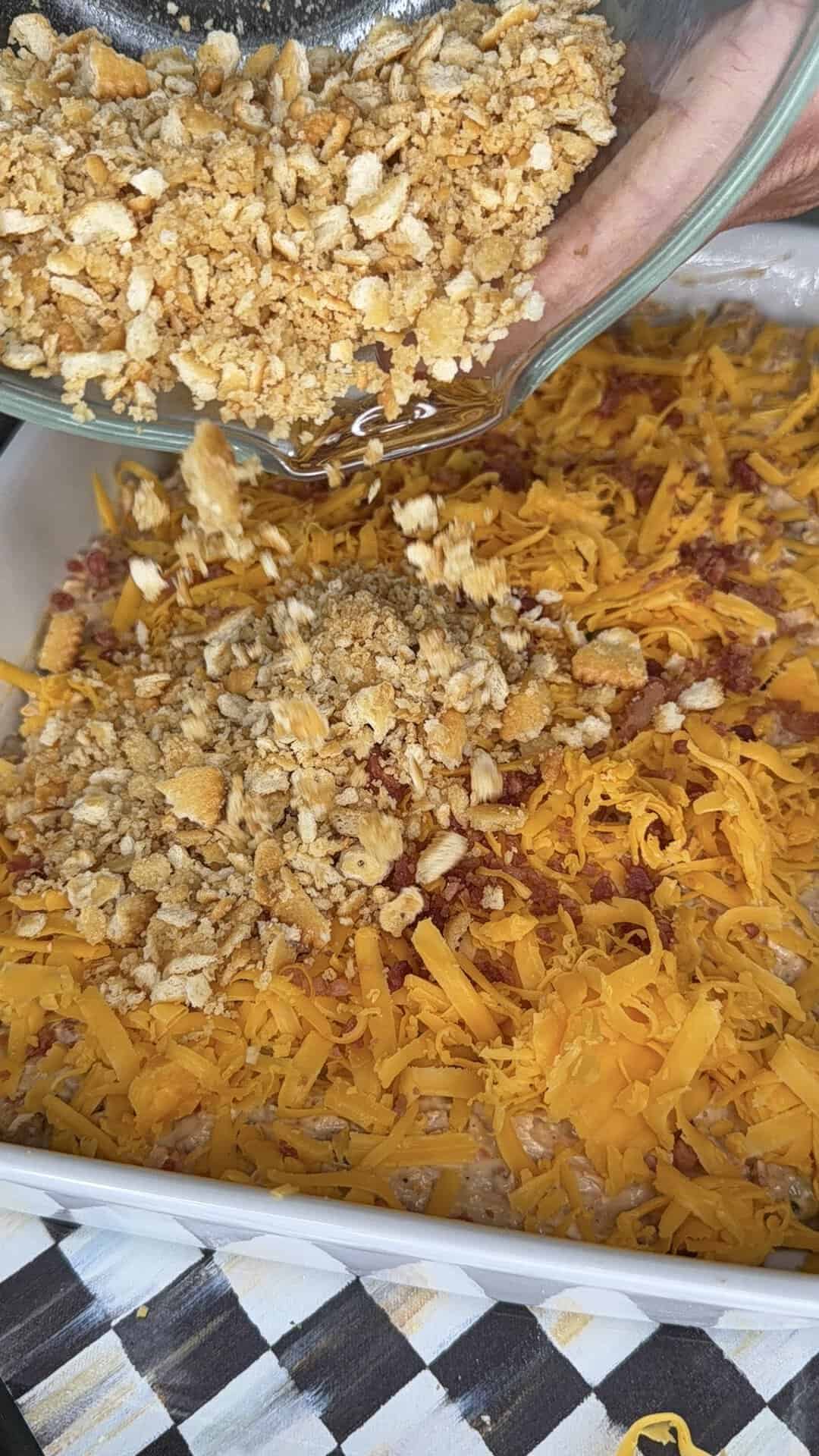 A hand pours crushed crackers from a glass bowl onto a casserole topped with shredded cheddar cheese and bacon in a white baking dish.