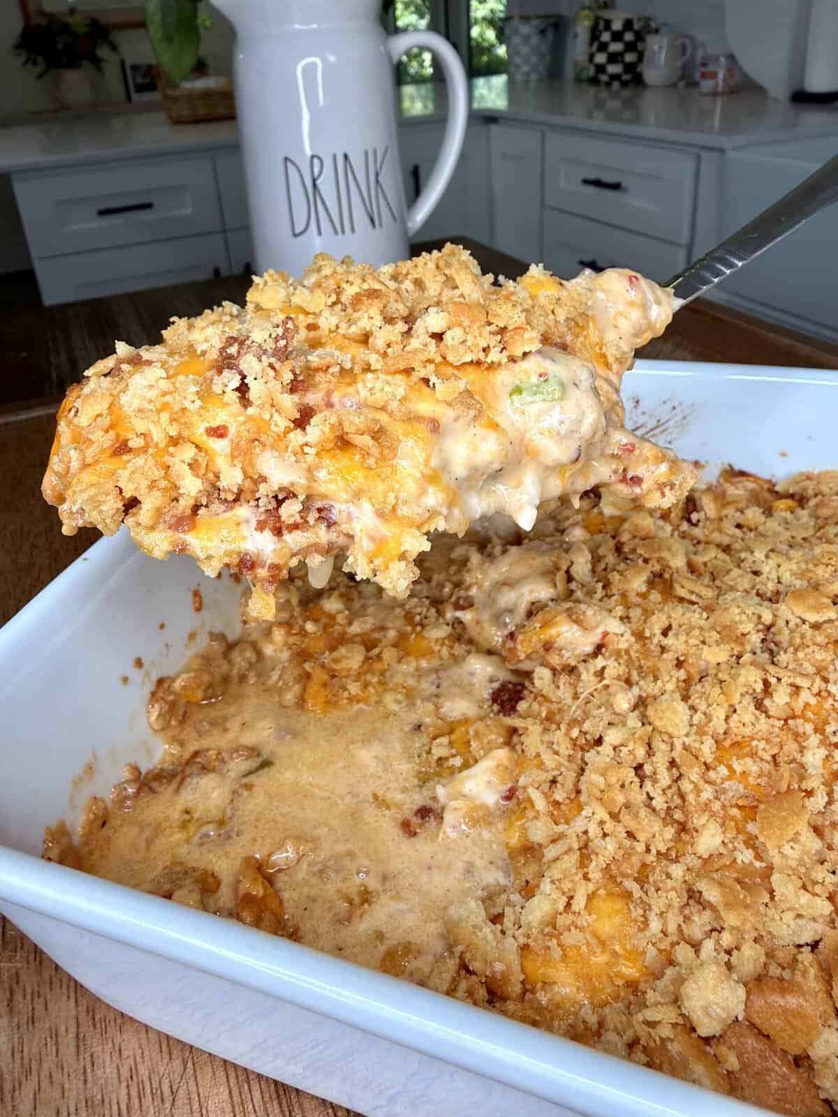 A close-up of a casserole dish filled with creamy casserole topped with a golden, crunchy crumb topping. A serving is being lifted out with a spatula in a kitchen setting.