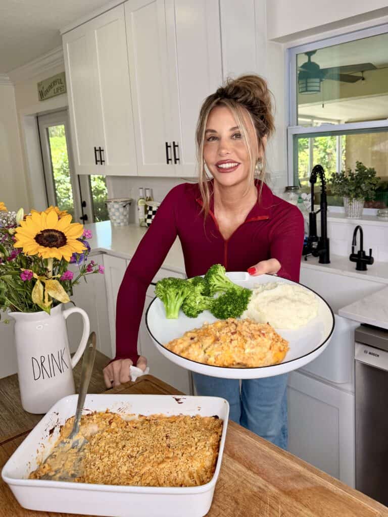 A smiling woman in a red top holds a plate with broccoli, mashed potatoes, and casserole in a bright kitchen, with a casserole dish and sunflowers on the counter.
