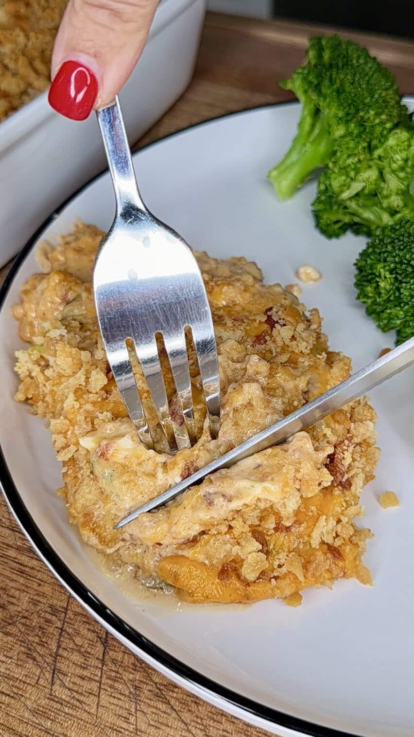 A hand with red nail polish holds a fork and knife, cutting into a cheesy casserole topped with a crumbly crust on a white plate, with broccoli on the side.