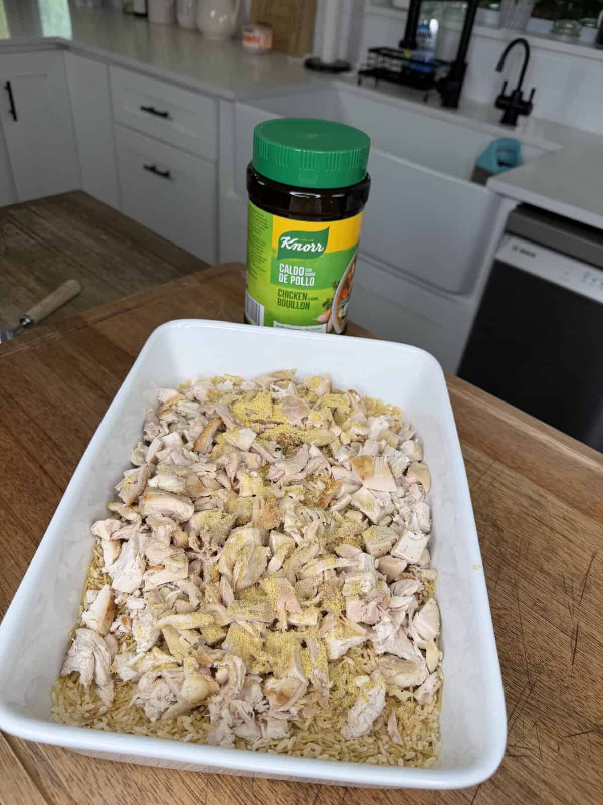 A white dish with uncooked rice and shredded chicken sits on a wooden counter. Chicken bouillon powder is sprinkled on top. A jar of Knorr chicken bouillon is behind the dish in a bright kitchen.