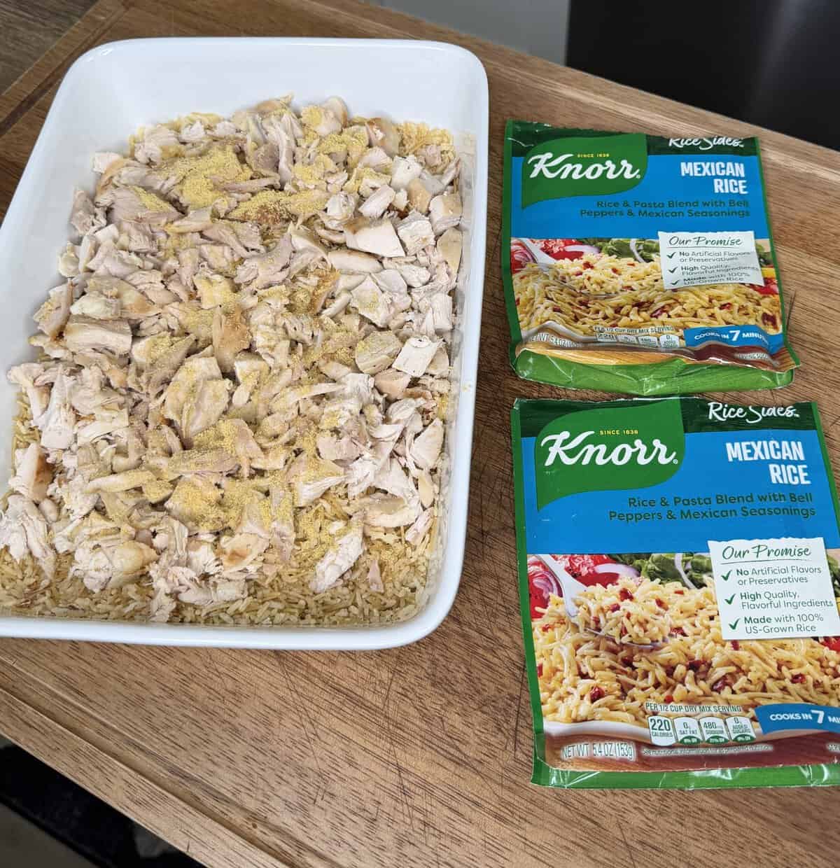 A white dish with cooked rice and shredded chicken, topped with seasoning, sits on a wooden surface. Two Knorr Mexican Rice seasoning packets are next to the dish.