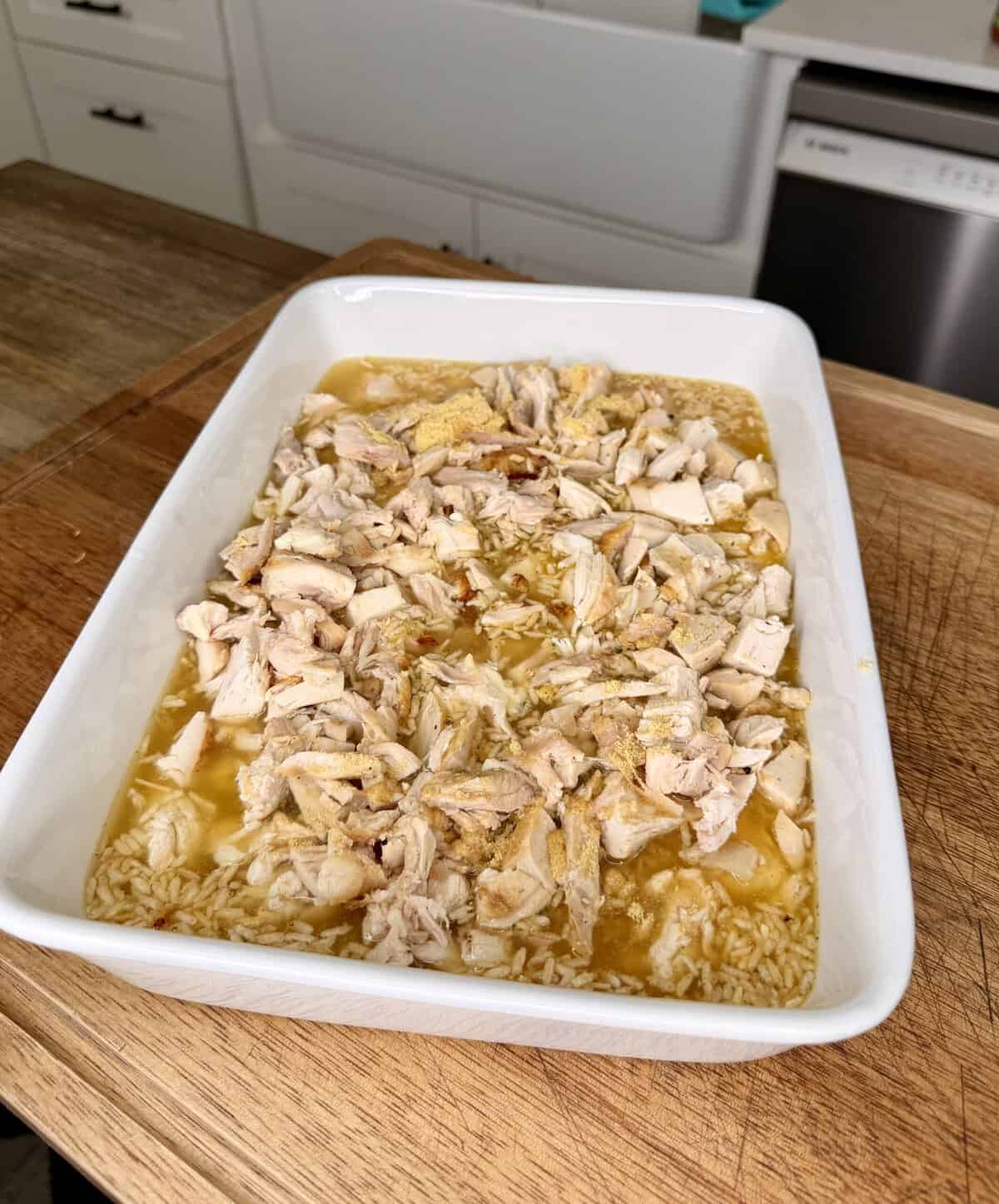 A white casserole dish filled with uncooked rice, broth, and chopped cooked chicken sits on a wooden countertop in a kitchen.