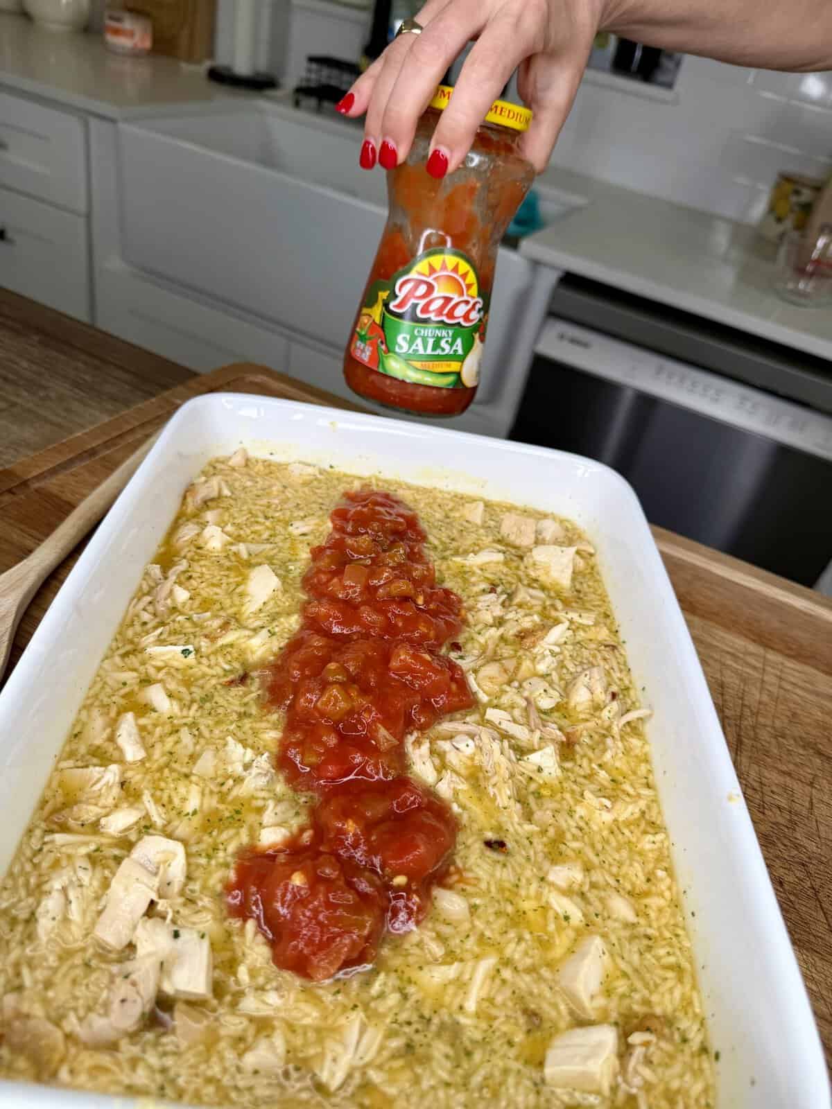 A hand holds a jar of Pace salsa above a casserole dish filled with rice and chicken, with salsa spread in a line down the center. The scene is set in a bright kitchen.