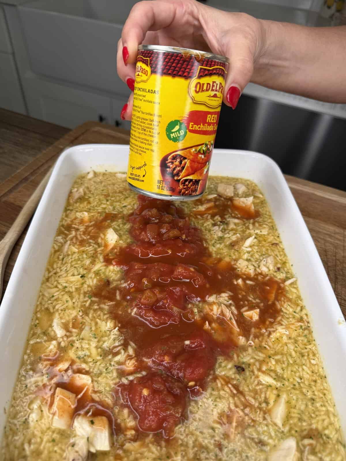 A hand pours Old El Paso red enchilada sauce over a baking dish filled with rice, chicken, and broth on a wooden counter.