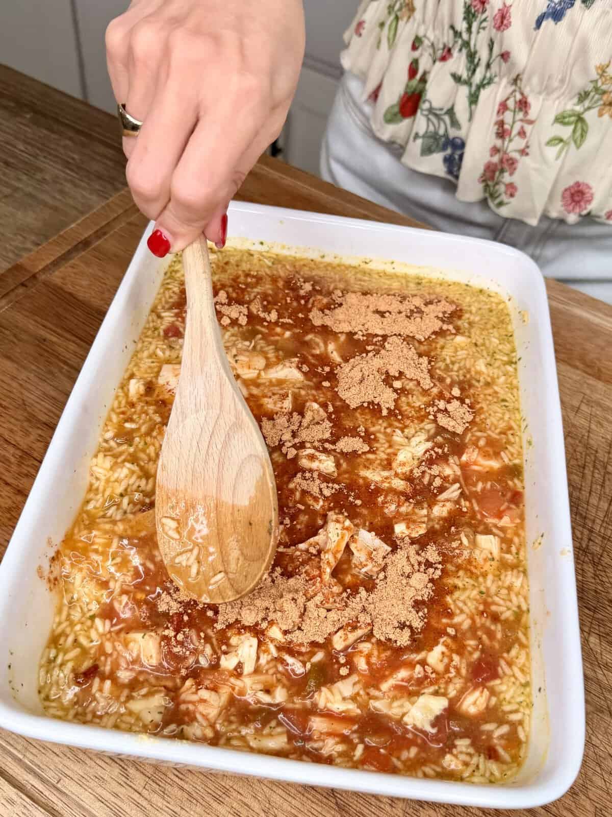 A hand stirs a baking dish filled with rice, liquid, chicken, and brown sugar using a wooden spoon. The person wears a floral top with red nail polish.