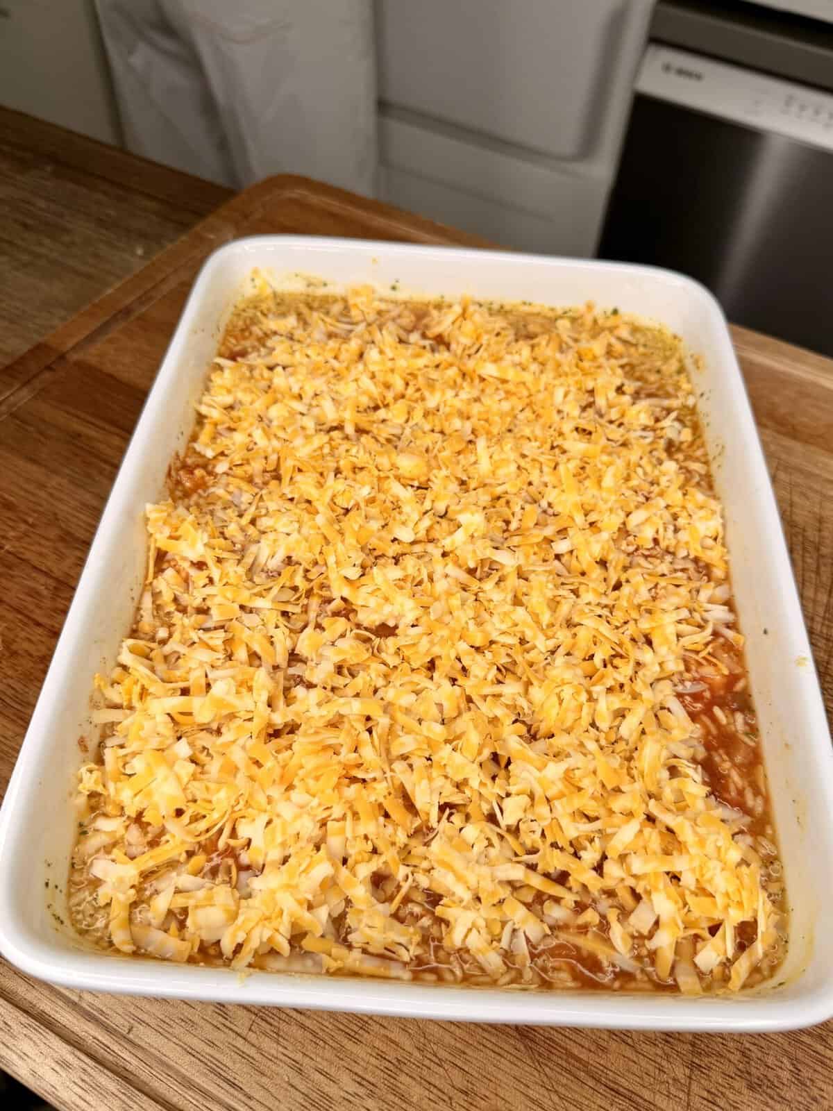 A rectangular white baking dish filled with a cheesy casserole sits on a wooden countertop. The top is covered with shredded yellow and white cheese, ready to be baked. Kitchen appliances are visible in the background.