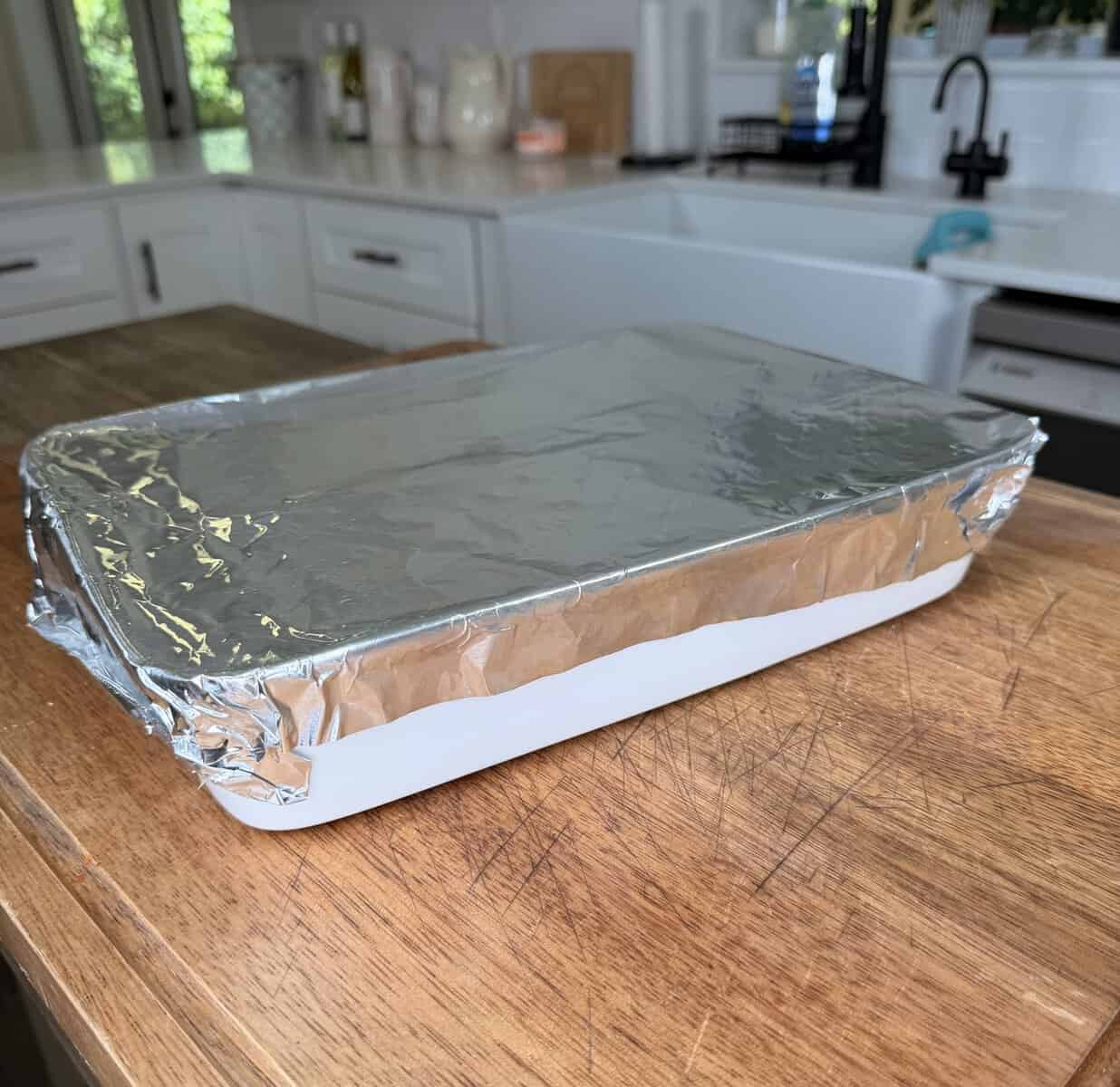 A rectangular white baking dish covered with aluminum foil sits on a wooden countertop in a bright kitchen.