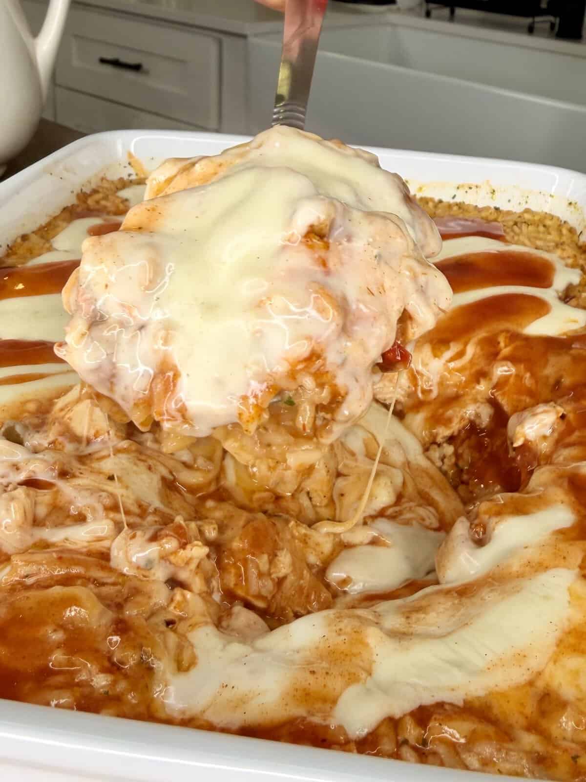 A close-up of a cheesy, saucy casserole in a white dish, with a serving being lifted out on a spatula. The casserole features melted cheese and a red sauce, and the kitchen counter is visible in the background.