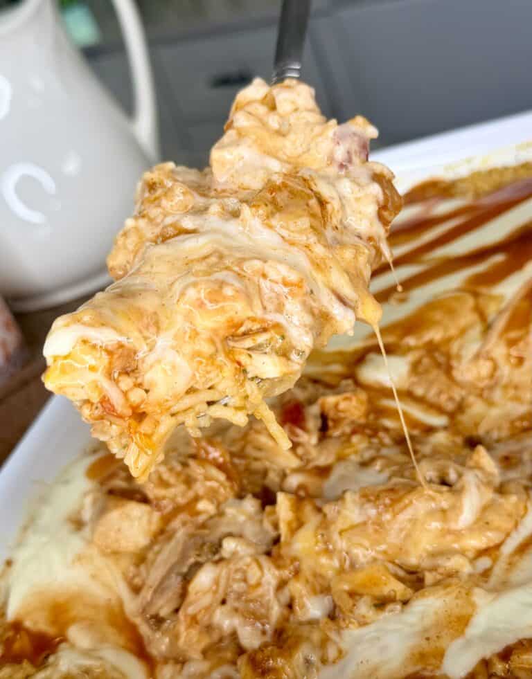 A close-up of a cheesy, saucy casserole with a spoon lifting a gooey serving, showing melted cheese and shredded meat inside a white dish.