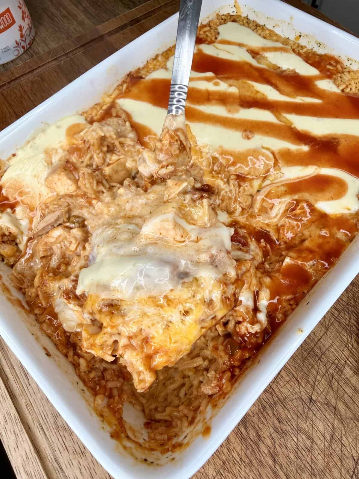 A casserole dish of cheesy baked rice topped with sauce, partially scooped out with a metal serving spoon on a wooden surface.