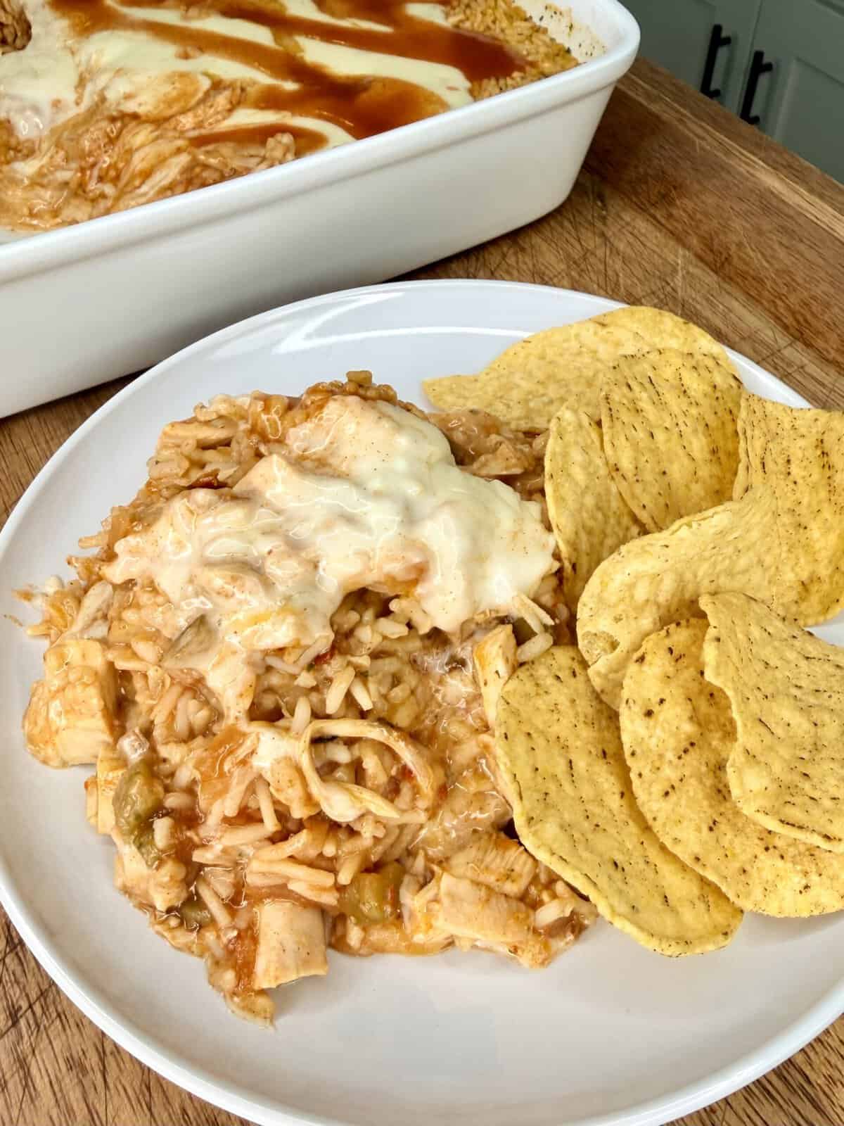 A plate with Mexican casserole topped with melted cheese, served with tortilla chips. A casserole dish sits in the background on a wooden surface.