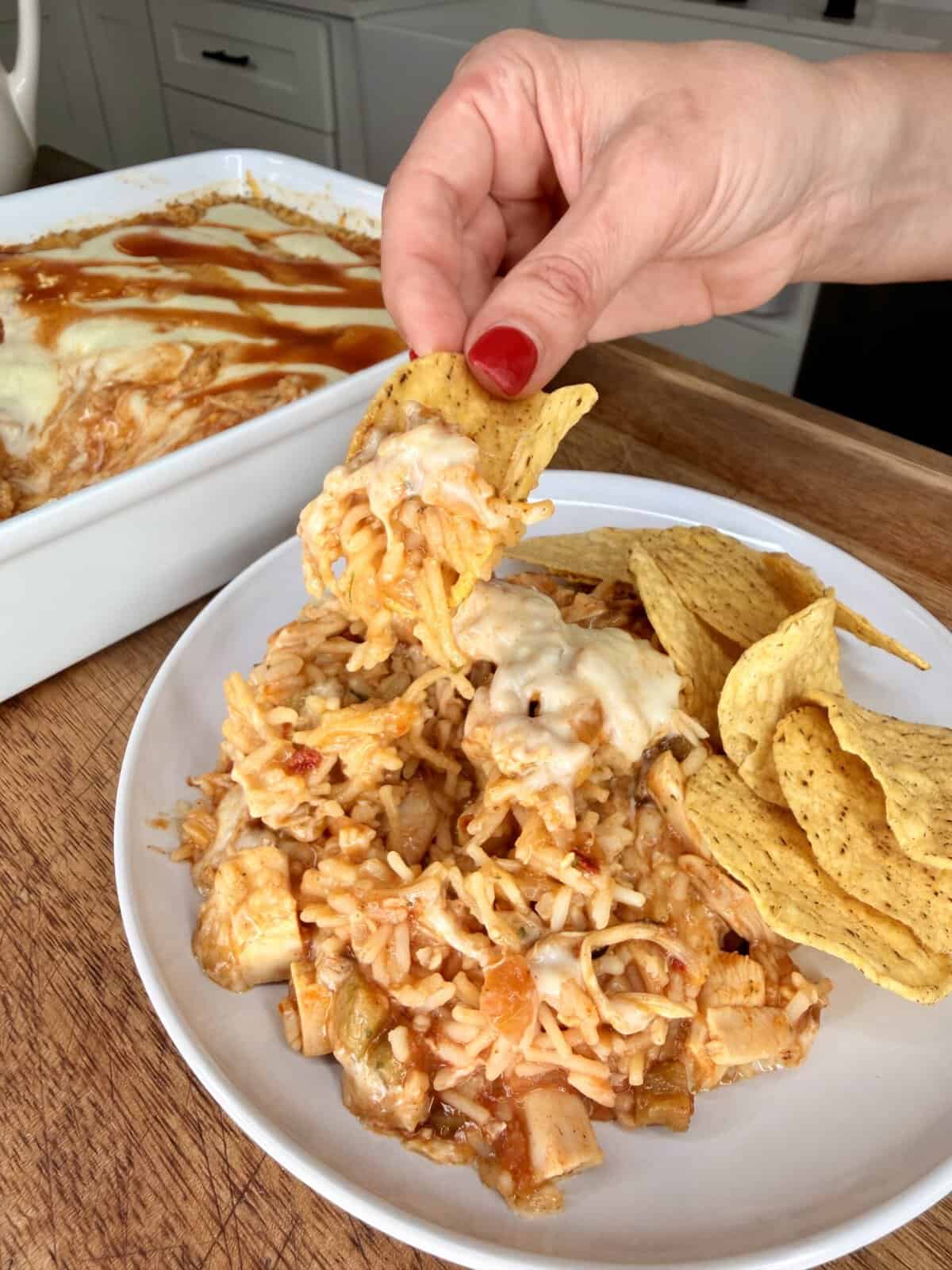 A hand scoops cheesy chicken casserole with a tortilla chip from a plate, with more chips and a casserole dish in the background on a wooden surface.