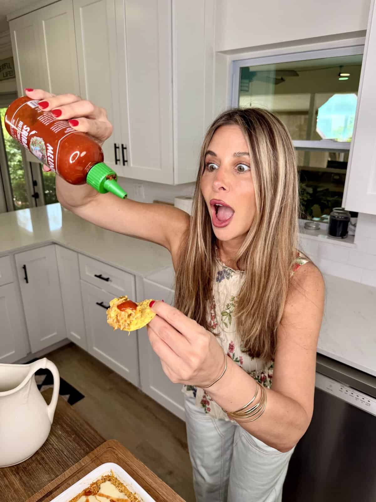 A woman in a kitchen enthusiastically squeezes sriracha sauce onto food, holding a surprised expression. She stands near a counter with an open casserole dish.