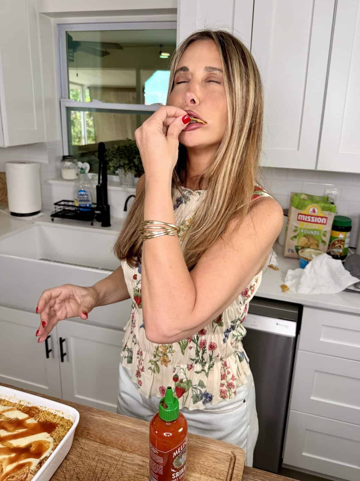 A woman stands in a kitchen, savoring food with her eyes closed and fingers near her mouth. A casserole dish and bottle of sriracha sauce are on the counter, with kitchen items and snack bags in the background.