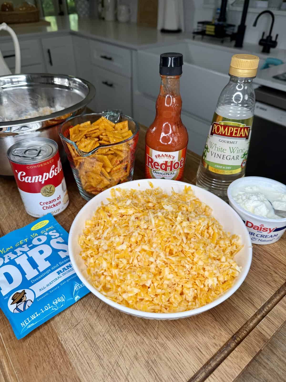 A kitchen counter displays shredded cheese in a bowl, a can of cream of chicken soup, ranch dip mix, Cheez-Its, hot sauce, white wine vinegar, sour cream, and a metal mixing bowl in the background.