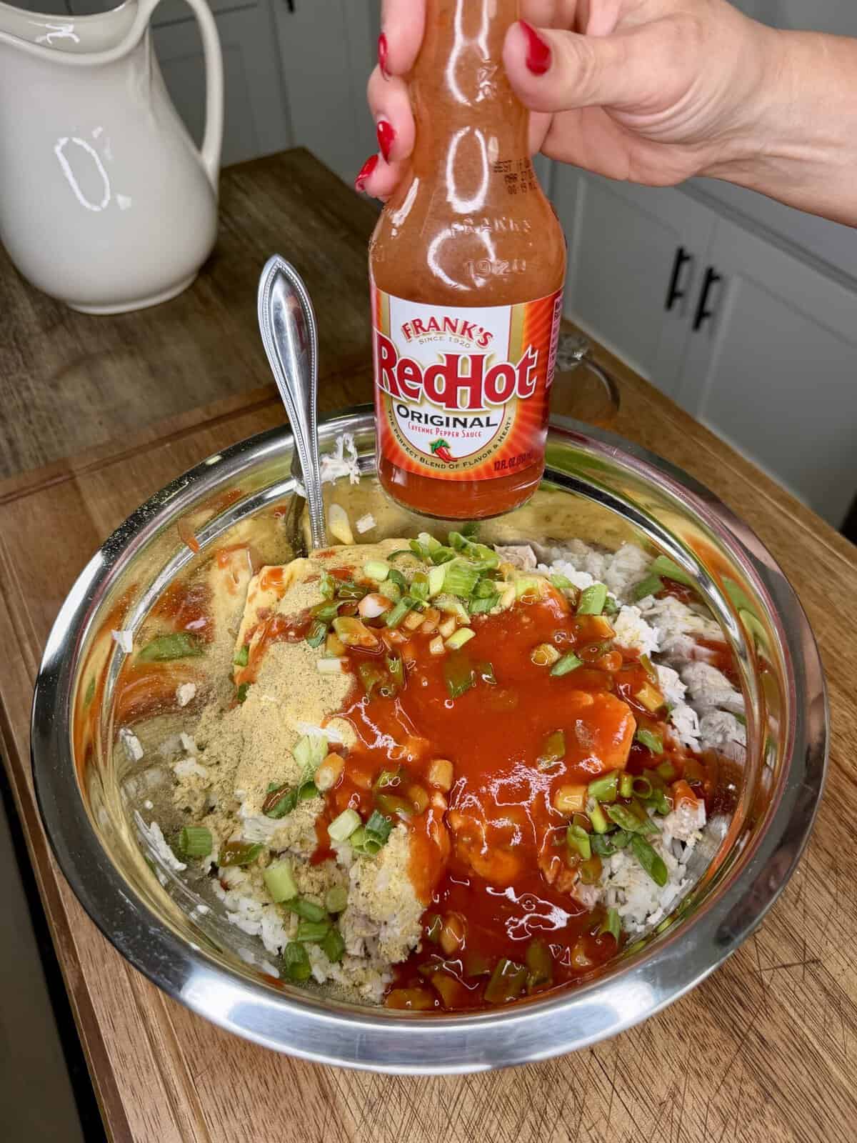 A hand holds a bottle of Franks RedHot sauce above a metal bowl filled with rice, chopped green onions, spices, and sauce on a wooden counter.