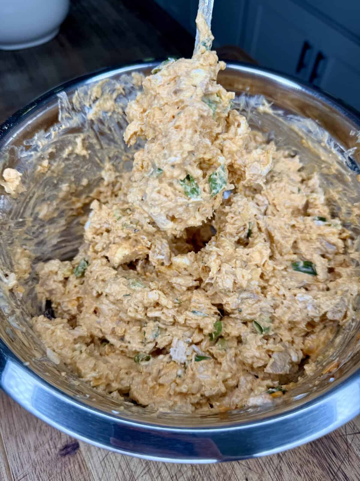 A metal bowl filled with a creamy, orange mixture containing chopped green herbs, being mixed with a spoon on a wooden countertop.