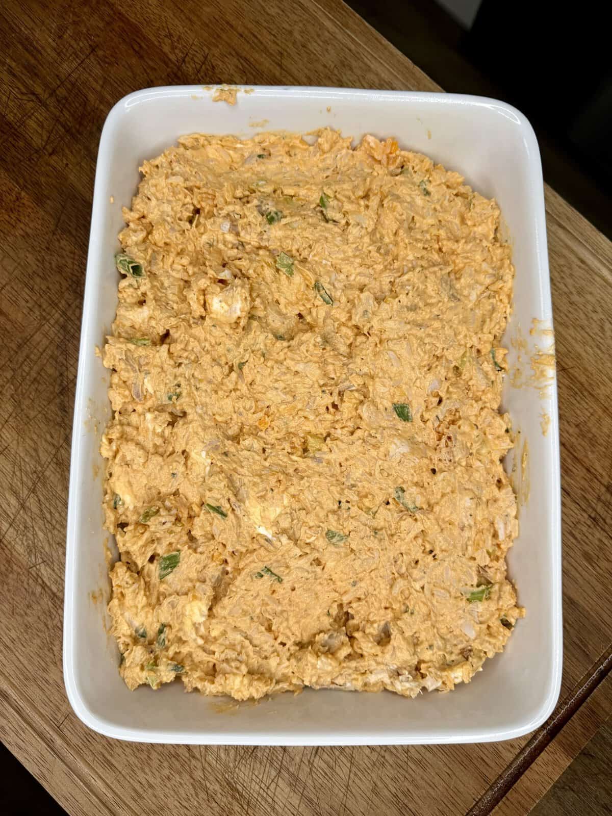 A rectangular white ceramic baking dish sits on a wooden cutting board, filled with a thick, orange-colored dip containing visible flecks of green herbs or scallions.
