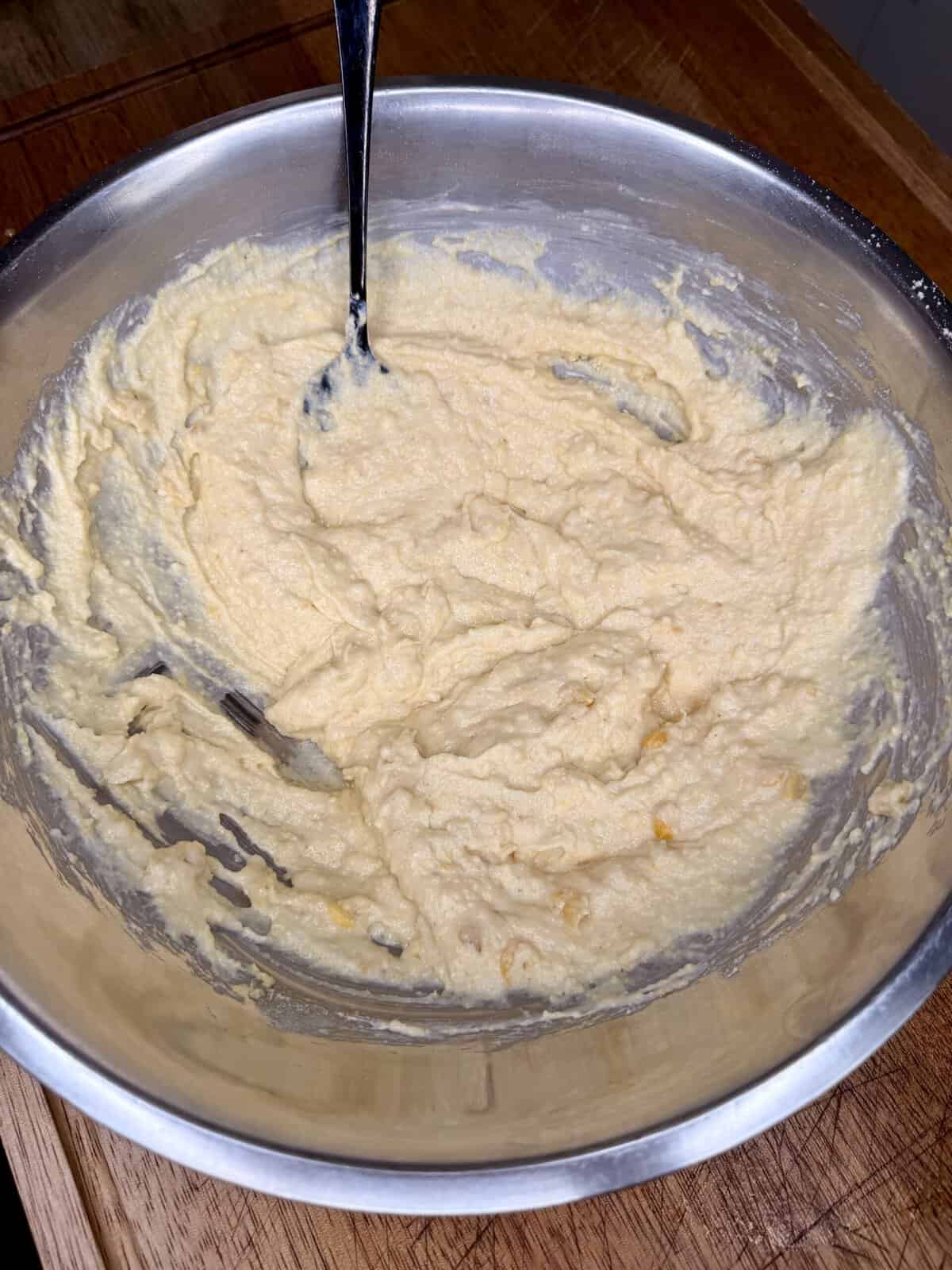 A metal bowl filled with thick, yellowish batter being mixed with a fork, placed on a wooden surface.