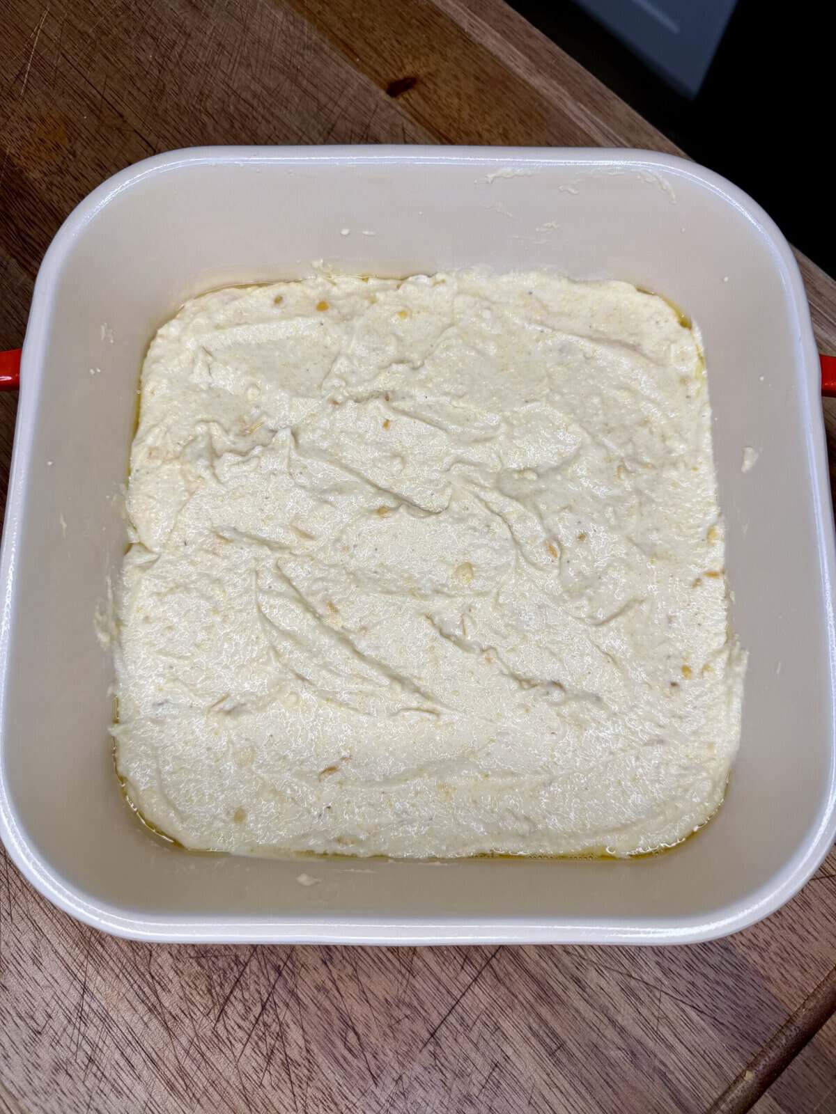 A square white dish filled with a smooth, creamy mixture sits on a wooden surface. The mixture appears pale yellow with some small lumps and a bit of oil along the edges.