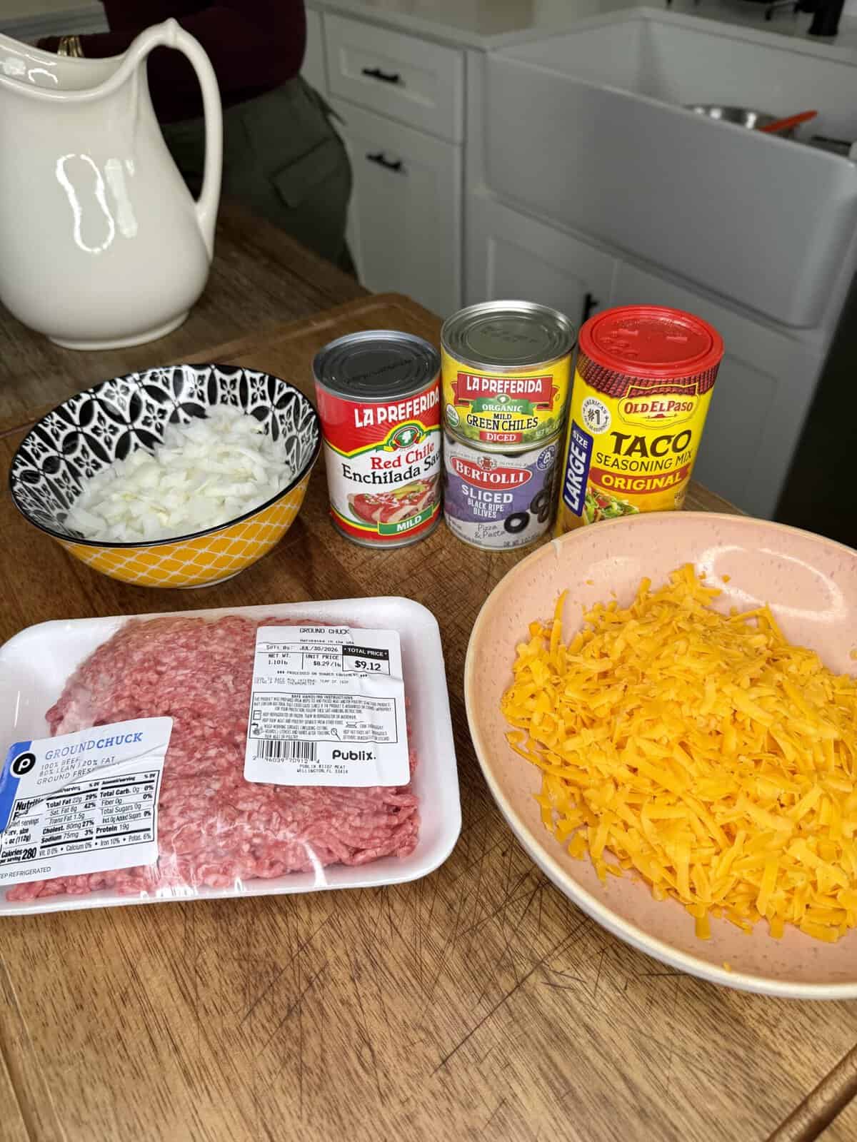 Chopped onions, ground chuck, shredded cheddar, canned enchilada sauce, diced green chiles, taco seasoning, and a bowl of rice sit on a kitchen counter next to a sink.
