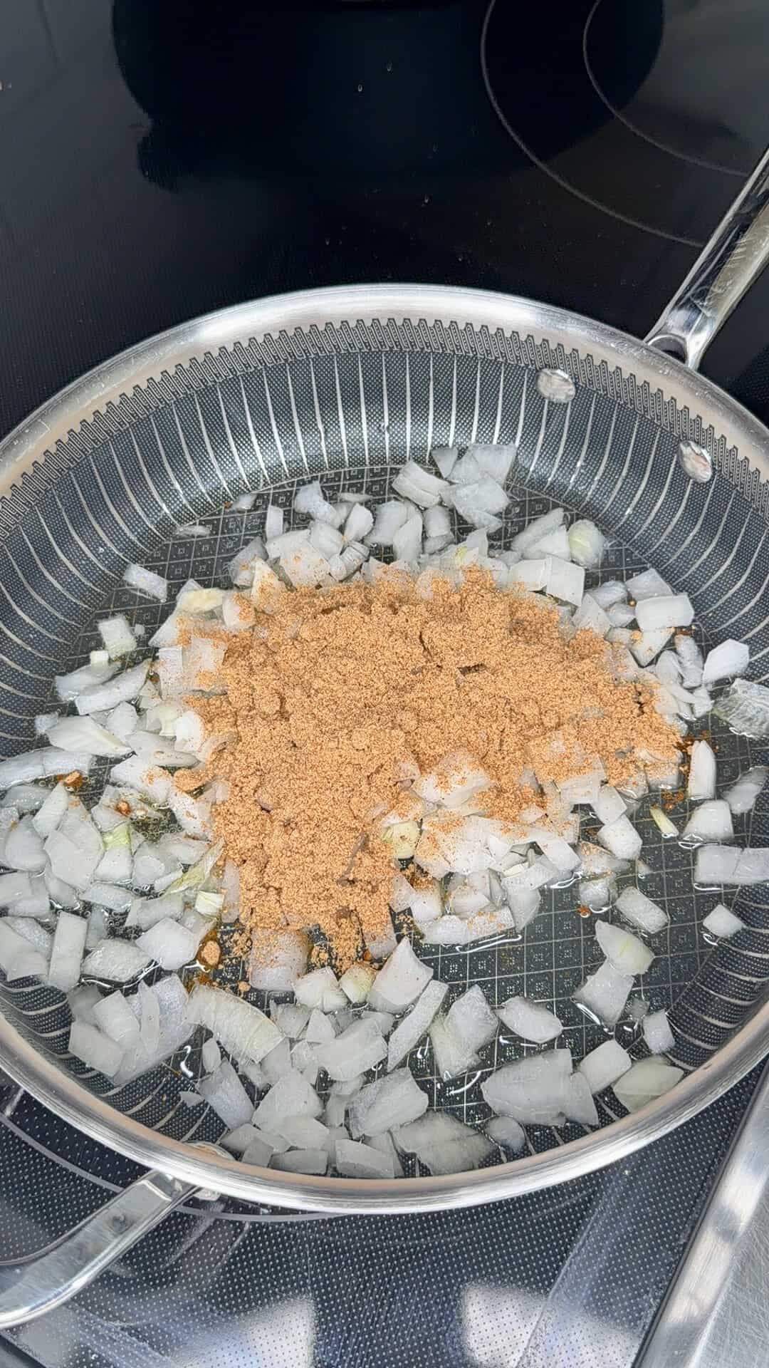 Chopped onions and a pile of brown seasoning mix are sautéing in a stainless steel pan on a stovetop.