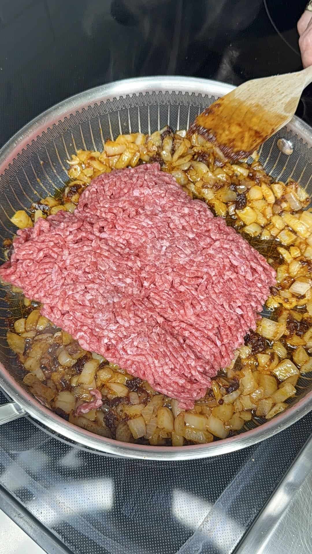 Raw ground beef is being added to a pan with sautéed diced onions. A wooden spatula is stirring the mixture on a stovetop, preparing to cook the beef.