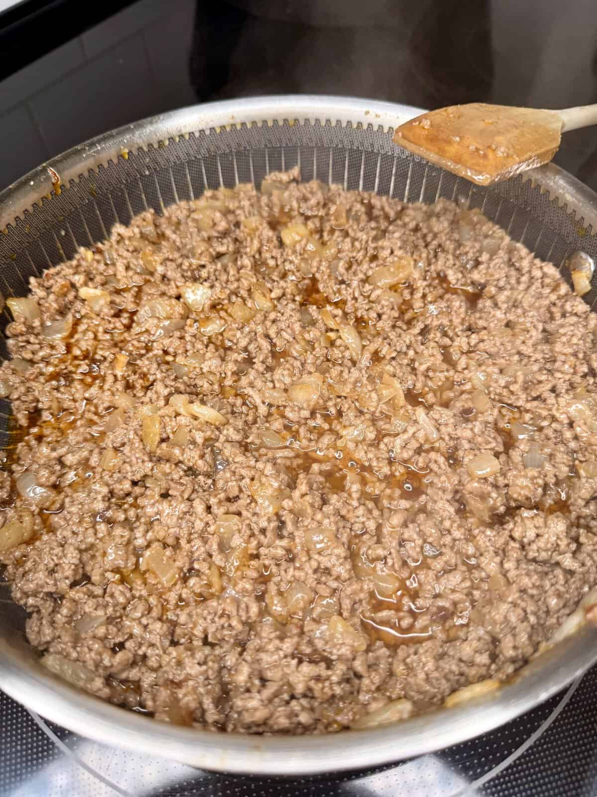 Ground beef and chopped onions are being cooked in a pan on a stovetop. A wooden spoon is resting on the edge of the pan. The beef is browning and there are visible juices.