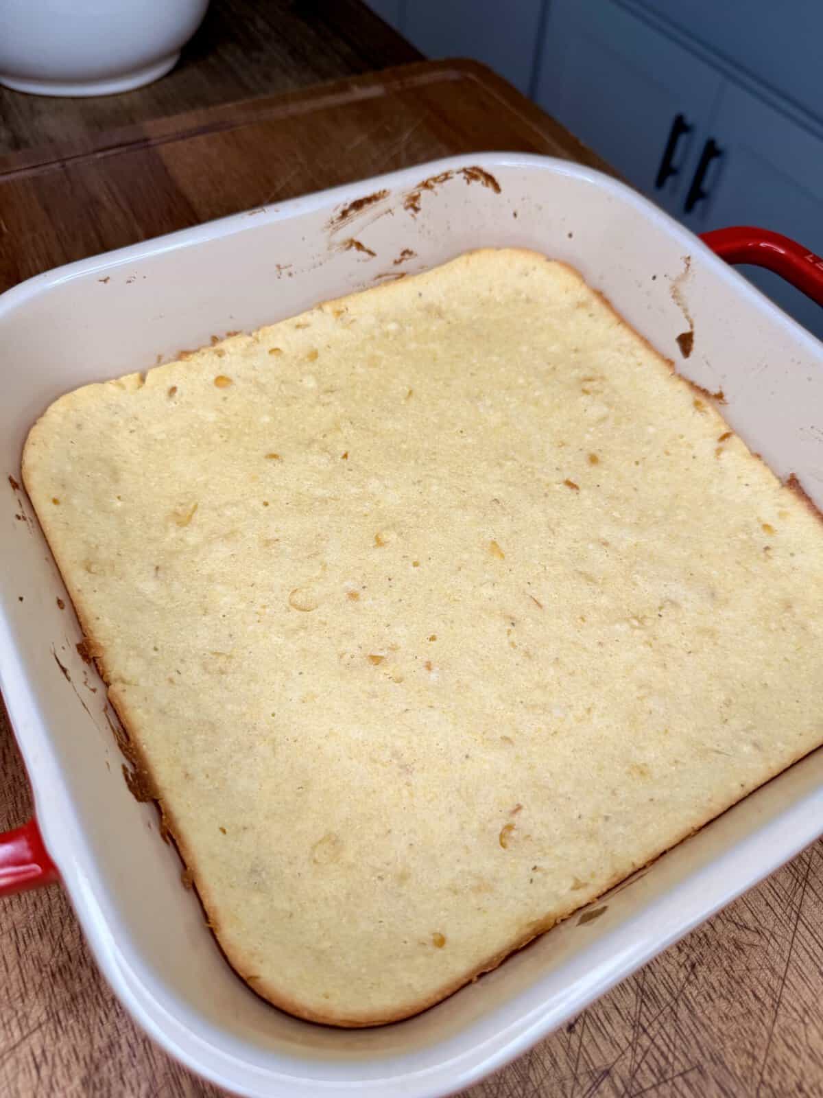 A square pan with golden-brown baked cornbread sits on a wooden surface. The edges are slightly darker, and the top appears smooth with a few small cracks. Red handles are visible on the side of the pan.