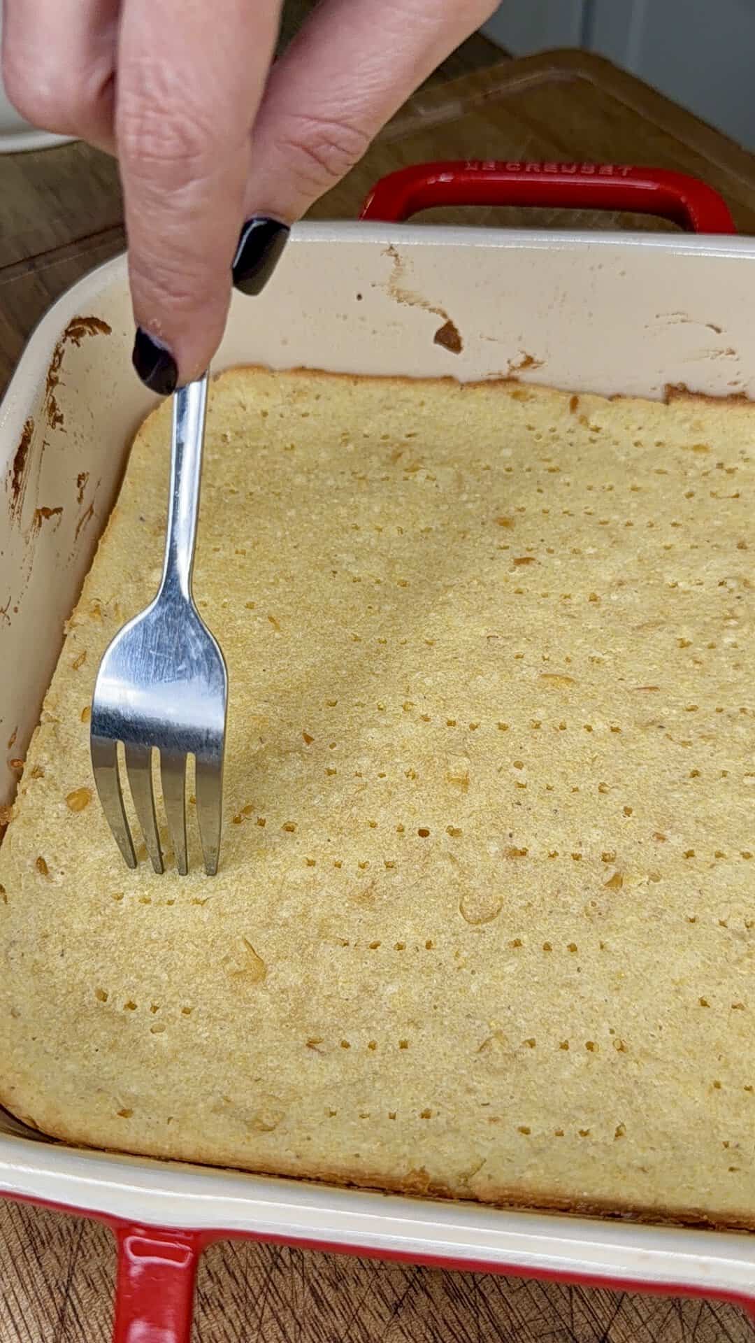 A hand with black nail polish uses a fork to poke holes into a golden, baked crust in a red rectangular baking dish on a wooden surface.