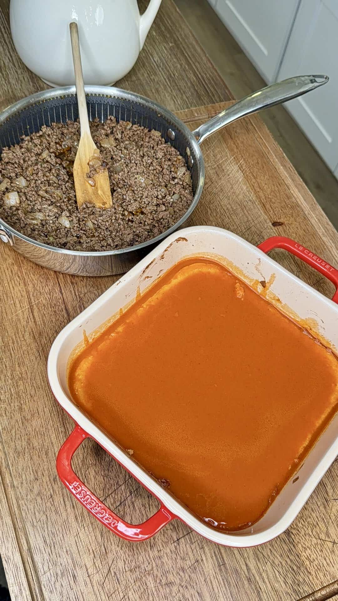 A skillet of cooked ground beef with onions sits beside a red dish filled with smooth orange sauce on a wooden countertop. A wooden spoon rests in the skillet.